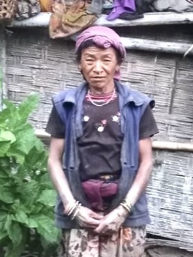 Mainsuba, wearing a purple headscarf and dark vest, standing in front of her weathered bamboo home in rural Nepal, hands clasped together